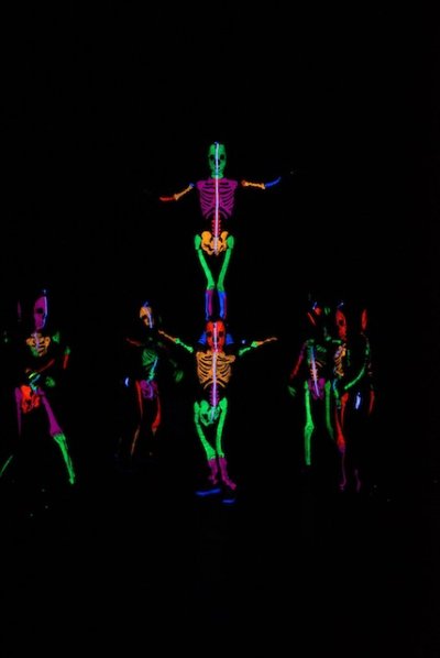 A group of student performers dressed in rainbow glow in the dark skeleton costume dance on stage. One pair is stacked, with one standing on anothers shoulders.