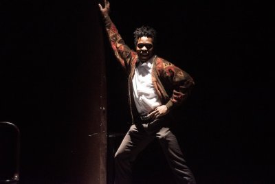 A student performer stands iin a disco pose on stage wearing a brown-red cardigan.