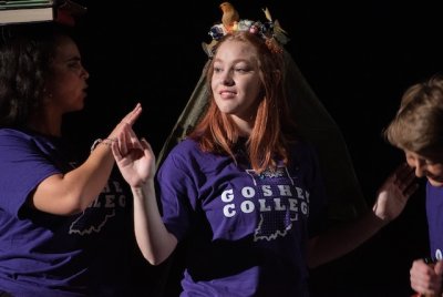 A female student performer representing "beauty" wears a purple GC shirt and a crown of flowers waves to the audience.