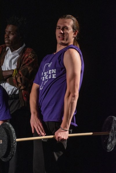 A student performer wearing a purple GC shirt with one sleeve raised to the shoulders is lifting weights, representing the character of Strength