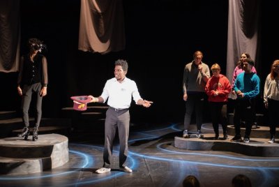 Male student performer wearing a white shirt and black pants addresses the audience while other characters ar eon stage unlit in the background.
