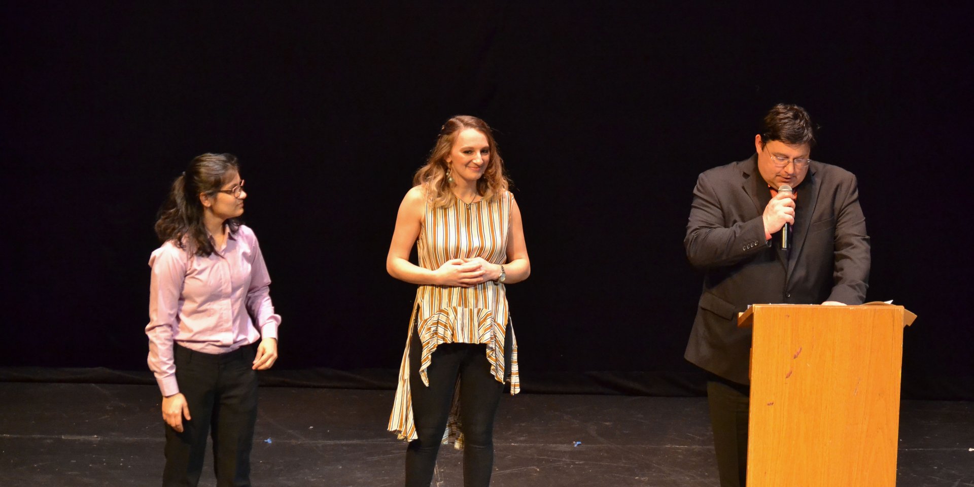 Male professor in front of the podium while two female students listen beside him.