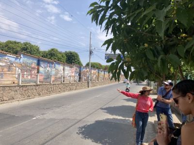 Along a main road, a brightly colored mural on one side and a group of people shaded by a tree on the other.